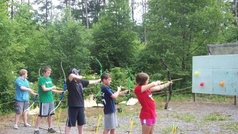 Youth shoot arrows at balloons pinned to a wooden board. 