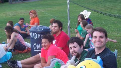 Counselors sit on the ground in front of a volleyball net. One counselor sits in a chair holding different stuffed animals. 