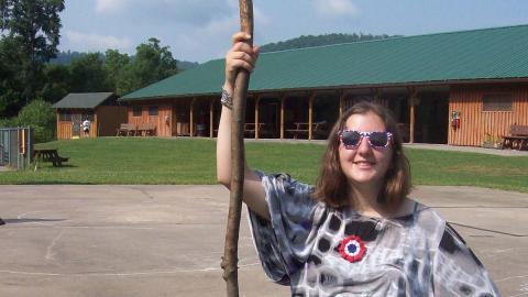 Youth holding a walking stick kneels on the ground with a white board and geocaching equipment in front. 