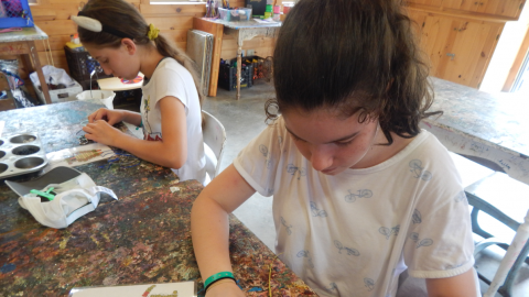 youth seated at a table strings beads according to a pattern in front of her to resemble winnie the pooh.Another youth in the background looks a pattern of an owl wearing a horned headband.