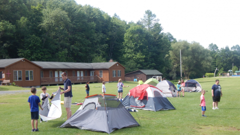 group of youth scattered among tents in the field. A counselor helps two youth holding a rain fly. 