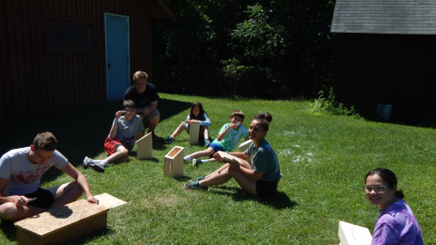 Youth sitting outside with woord working projects in the sun as counselor demonstrates burning wood through a magnifying glass.
