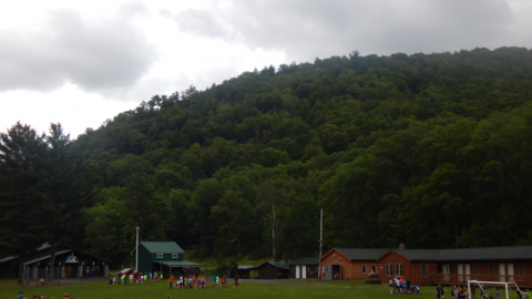 Wide shot of camp showing forest in distance of buildings with campers in foreground.