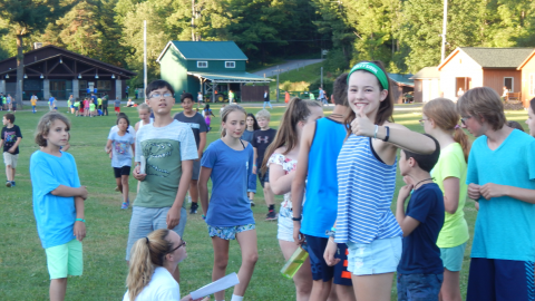 Group of campers around a seated counselor in the field during a camp-wide game. One youth wearing a camp bandanna smiles and gives the camera a thumbs up.