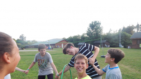 campers standing in a circle smiling as one camper tries to duck through a hula hoop on his arm.