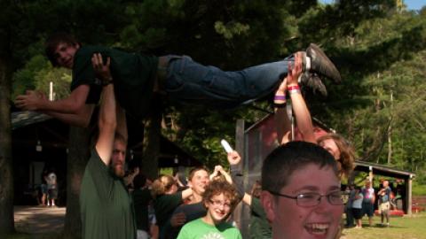 campers coming toward the camera through a funneled line of counselors. The closest counselor is raised up over their heads by two tall counselors and smiling at the kids below.