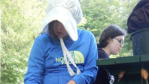 Camper wearing a colonial hat shaves the bark of off a stick during a living history class. 