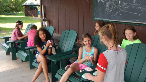 Group of youth seated in and behind Adirondack chairs on the front porch.