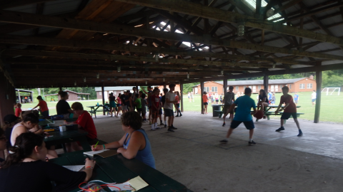 distance view of rec hall with campers playing 4-square and seated at picnic tables.