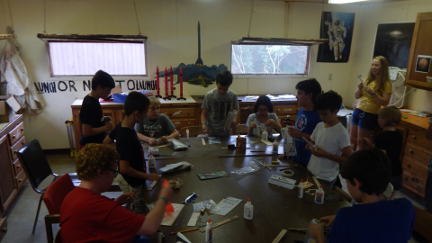 youth seated around the table in various stages of building model rockets as counselor looks on with hot glue gun.