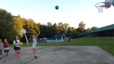 youth lined up on the basketball court during a game of horse. Two youth hold basketballs. One has just made a shot and has arms extended toward the hoop with ball mid-air.