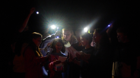 cabin group gathered together in the dark with flashlights to help them sing lyrics written on papers they are holding.