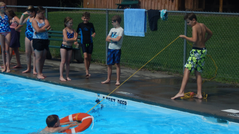 camper pulling another camper to pool side with a rescue ring while others camp watch from the pool deck.