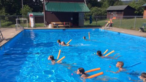 Youth swimming in the pool race across with pool noodles.