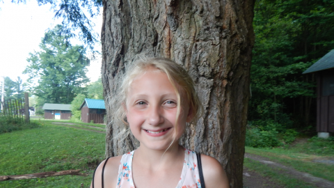 Campers stands smiling in front of a tree with cabins in the background.