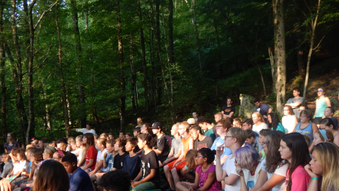 campers seated on benches in a wooded area watching a performance by a platoon group in front.