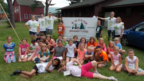 Young campers sitting on ground in front of Camp Shankitunk sign, two laying on the ground in front. Counselors standing in back