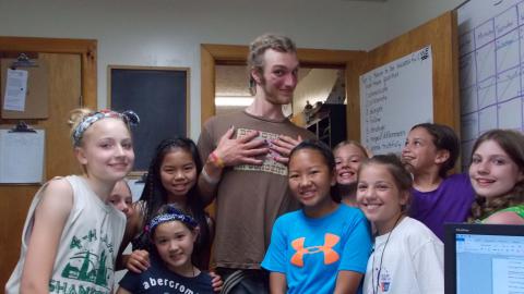 group of young ladies pose posing with tall counselor they have just "made over." He has pink/purple nails, braided hair, and facial make-up.