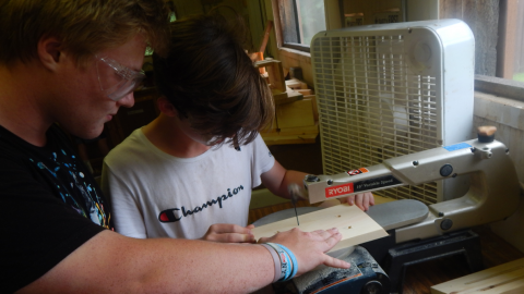 counselor works with one camper at the drill press carefully saw a long slit in the center of a board. Both are wearing safety goggles.